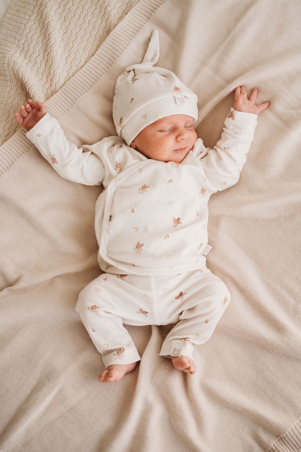 Een vredige pasgeboren baby in een Muts Cocoa-outfit en een hoogwaardige bijpassende Yumi Baby-muts, heerlijk slapend op een zachte, neutraal getinte deken.