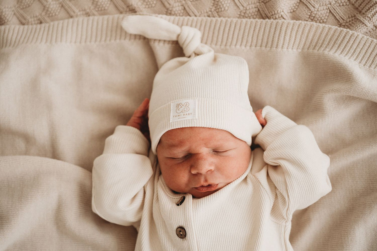 Een pasgeboren baby in een knus wit Boxpakje Ivory Dreams van hoogwaardig katoen en bijpassend mutsje slaapt vredig op een zachte, gestructureerde deken.