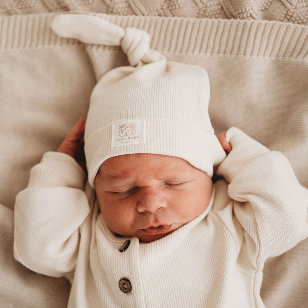 Een zachte pasgeboren baby, gekleed in een gezellig wit onesie en bijpassend Muts Ivory Dreams mutsje van Yumi Baby, ligt diep te slapen op een zacht beige.