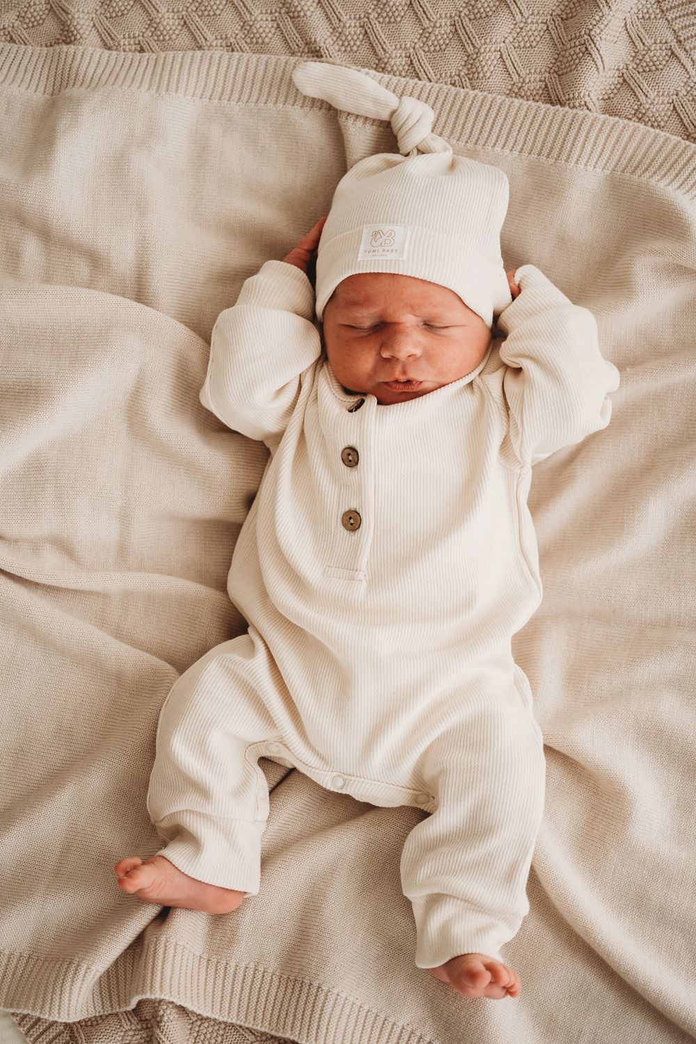 Een zachte pasgeboren baby, gekleed in een gezellig wit onesie en bijpassend Muts Ivory Dreams mutsje van Yumi Baby, ligt diep te slapen op een zacht beige.