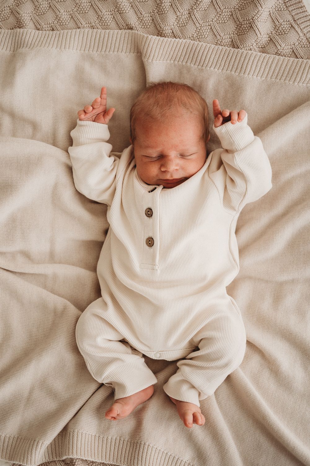 Een pasgeboren baby in een knus wit Boxpakje Ivory Dreams van hoogwaardig katoen en bijpassend mutsje slaapt vredig op een zachte, gestructureerde deken.