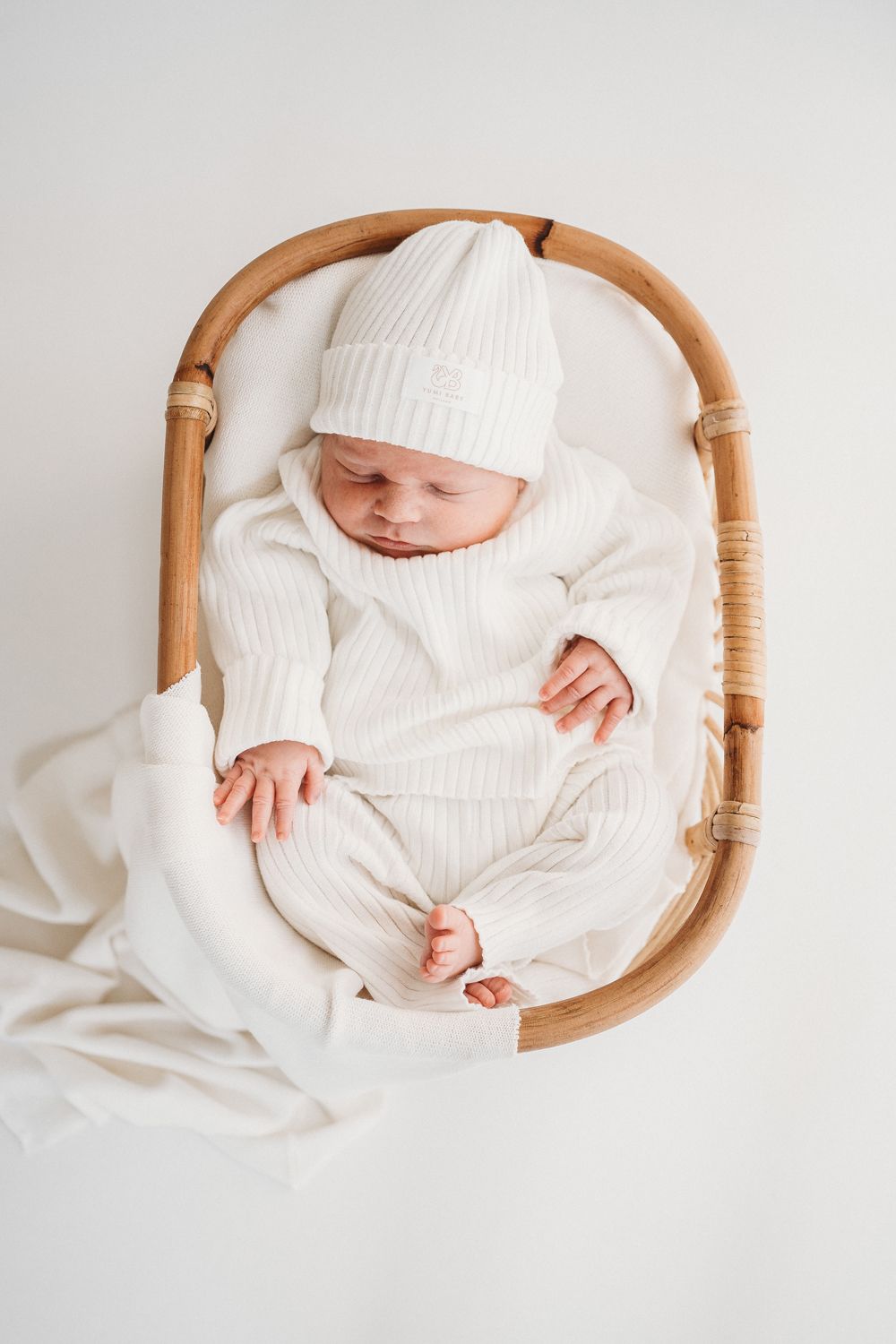 Een serene pasgeboren baby, gekleed in het wit, slaapt vredig in een knus, ovaalvormig rieten wiegje, dat een nieuw begin en pure onschuld symboliseert. De setting wordt versterkt door het comfortabele Yumi Baby Knitted broekje met parelmoer voor de baby.