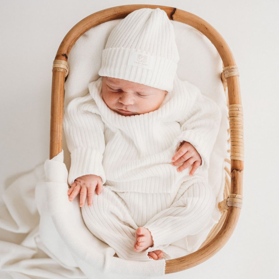 Een serene pasgeboren baby, gekleed in een Yumi Baby Gebreide Trui met Parelmoer, comfortabel genesteld in een geweven wiegje met een zachte deken, die vredige onschuld belichaamt.