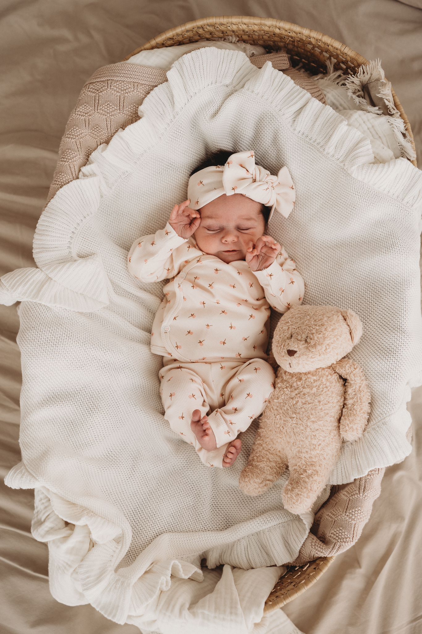 Een vredige pasgeboren baby in een knus outfitje van organisch katoen slaapt heerlijk in een geweven mandje, geknuffeld door zachte dekentjes met een Haarband Tiny Tulip van Yumi Baby metgezel aan hun zijde.