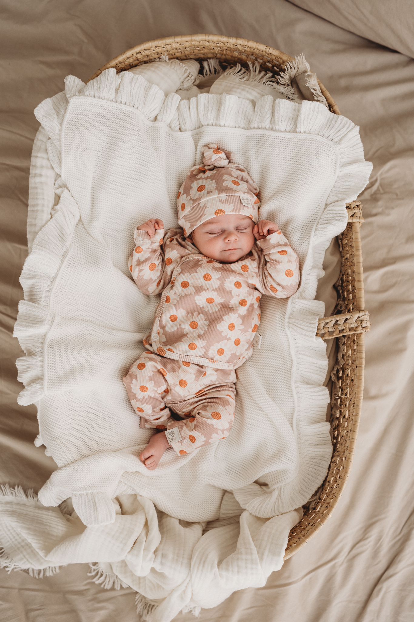 Een vredige pasgeboren baby slaapt heerlijk in een mandje, gekleed in het schattige Yumi Baby Broekje Daisy Delight met bijpassend mutsje, genesteld in een knus dekentje.