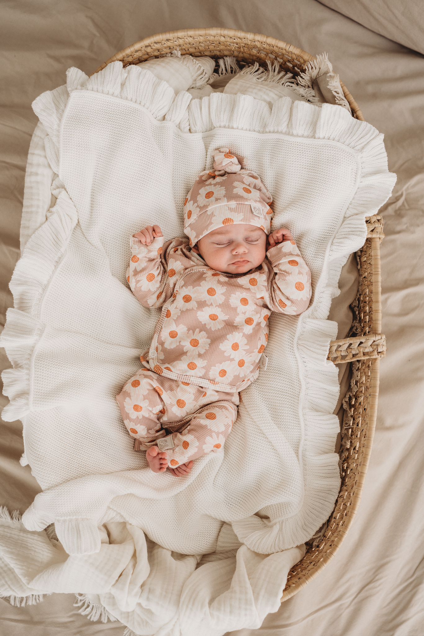 Een serene newborn slaapt vredig in een knus rieten mandje, gekleed in een schattige onesie met een bijpassend Yumi Baby pasgeboren babymutsje, omringd door een zachte, crèmekleurige deken.