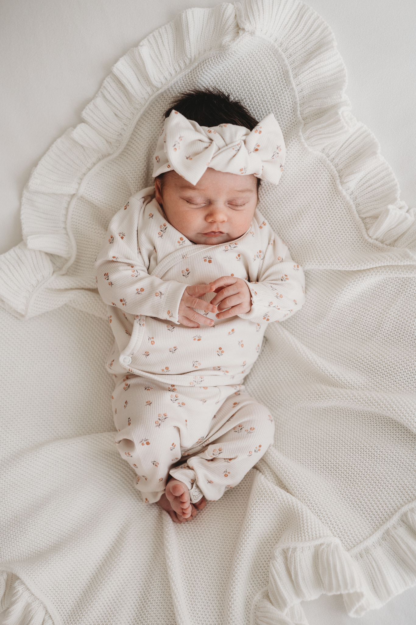 Serene slaap: een schattige baby gekleed in een gebloemde outfit met een bijpassende haarband Peach Blossom van Yumi Baby slaapt vredig op een zachte, gegolfde deken.