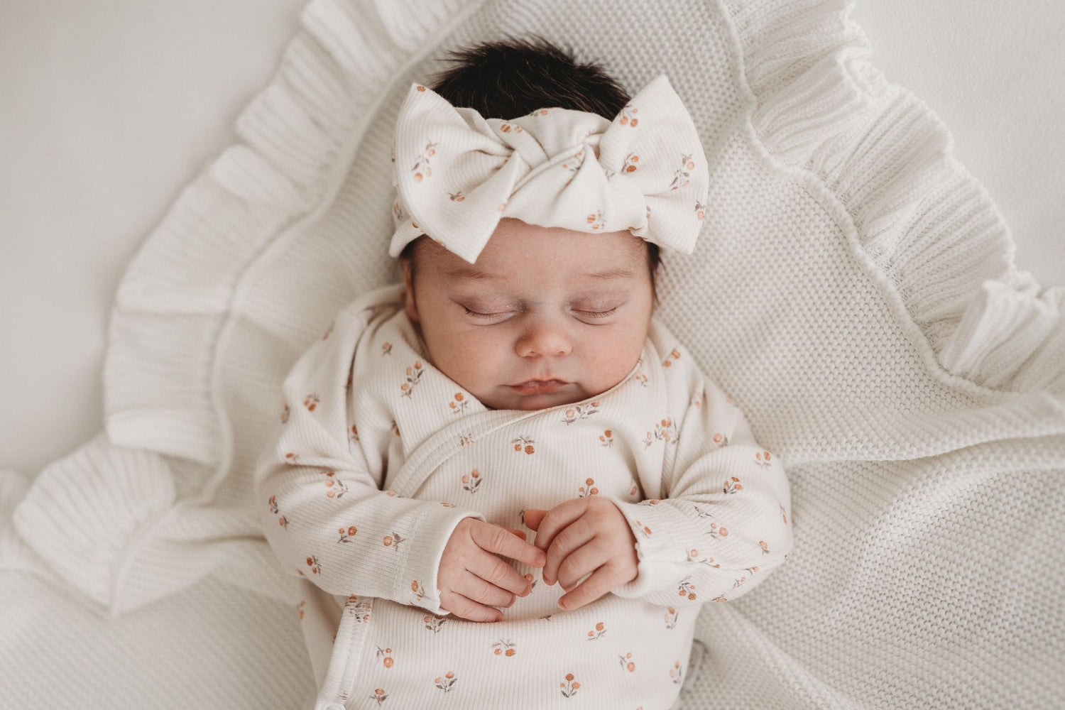 Serene slaap: een schattige baby gekleed in een gebloemde outfit met een bijpassende haarband Peach Blossom van Yumi Baby slaapt vredig op een zachte, gegolfde deken.
