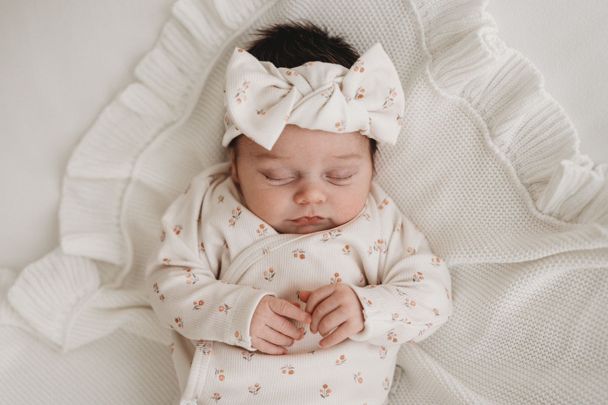 Een tevreden baby slaapt vredig, gekleed in een gezellig Yumi Baby overslagshirt perzikbloesem met bijpassend mutsje, genesteld in een zachte, witte deken.