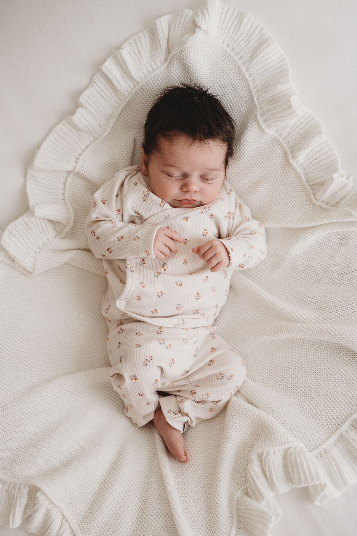 Een vredige pasgeboren baby die slaapt, gekleed in een schattig bedrukt Yumi Baby Broekje Peach Blossom onesie en bijpassend hoedje, liggend op een zacht wit oppervlak met de armen zachtjes omhoog bij het gezicht.