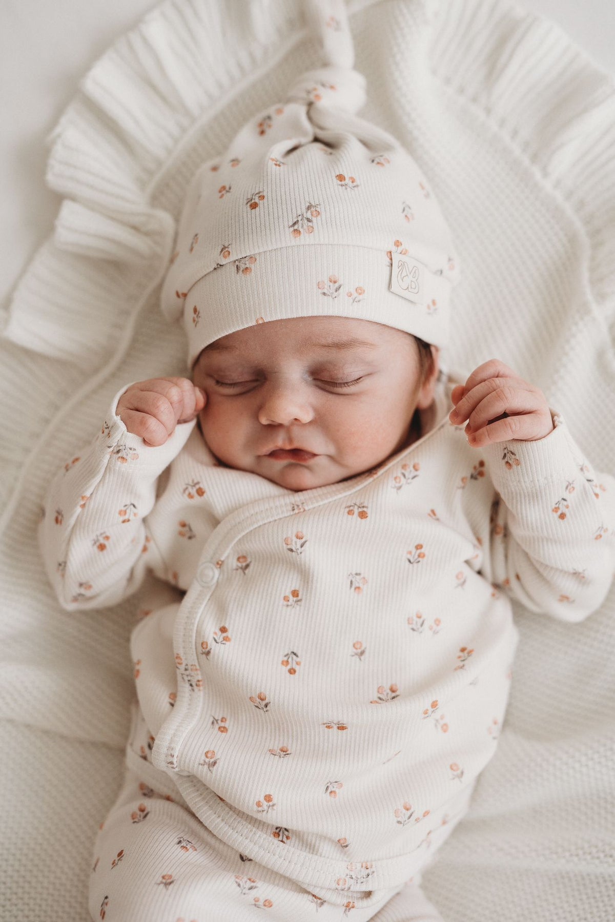 Een vredige pasgeboren baby die slaapt, gekleed in een schattig bedrukt Yumi Baby Broekje Peach Blossom onesie en bijpassend hoedje, liggend op een zacht wit oppervlak met de armen zachtjes omhoog bij het gezicht.