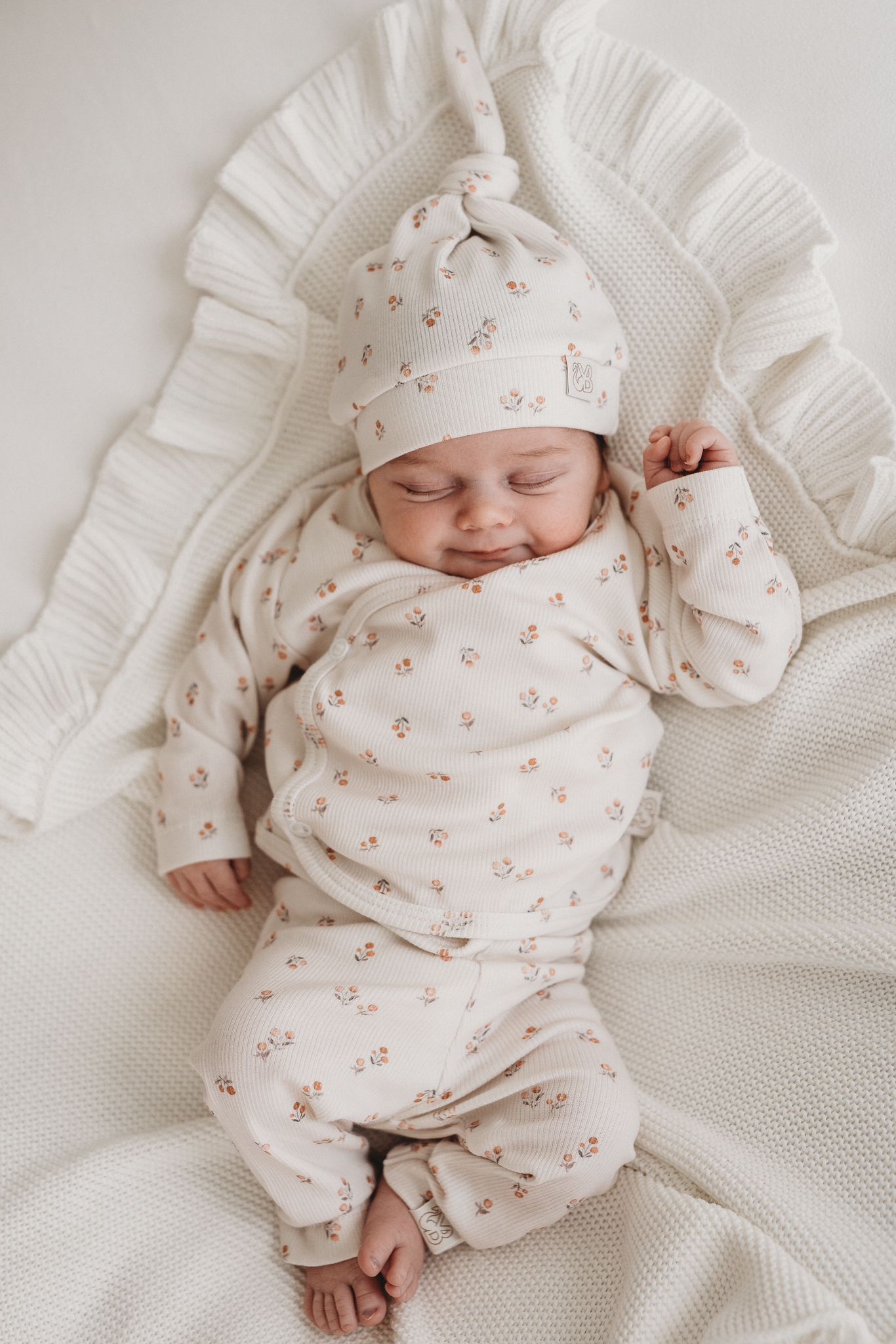 Een tevreden baby slaapt vredig, gekleed in een gezellig Yumi Baby overslagshirt perzikbloesem met bijpassend mutsje, genesteld in een zachte, witte deken.