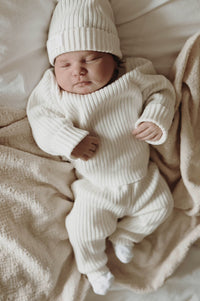 Een vredige pasgeboren baby, gekleed in een Yumi Baby Knitted Broekje Pearl Whisper en muts, rustend op een zachte, gekreukelde deken met een comfortabele pasvorm.