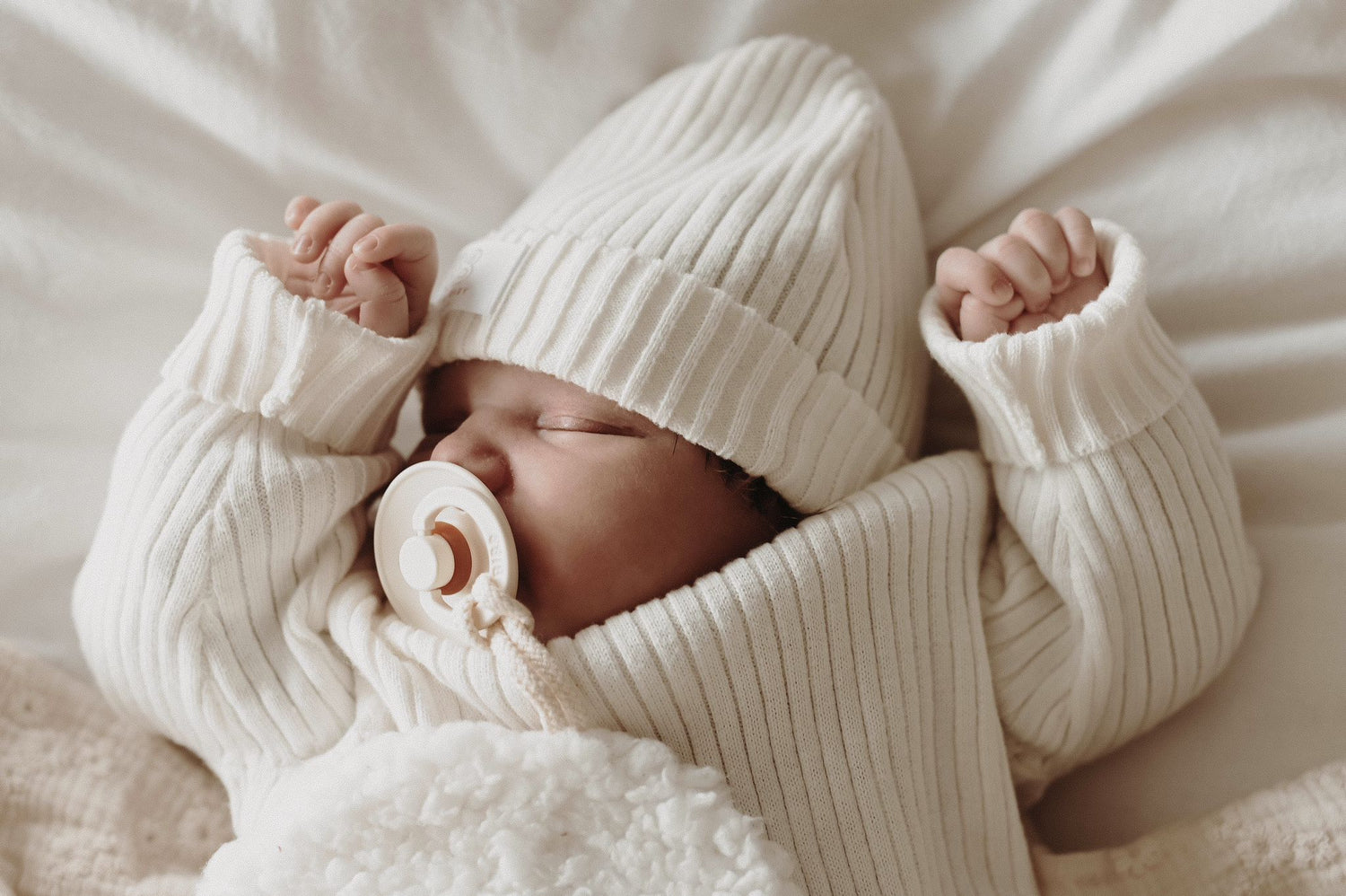 Een vredige pasgeboren baby, gekleed in witte kleding, lag comfortabel in een klein rotan wiegje, met een zachte deken en een delicate Yumi Baby Knitted Muts Pearl Whisper 0-3 mnd zachtjes gedrapeerd over de zijkant.