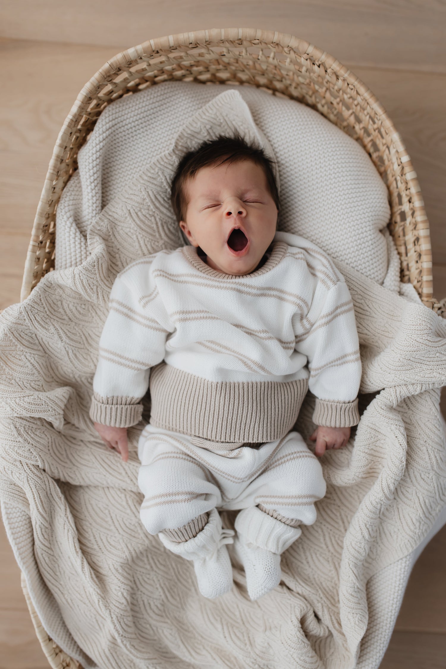 Een vredige pasgeboren baby gaapt terwijl hij comfortabel in een geweven wiegje ligt, gewikkeld in een zachte 100% Yumi Baby gebreide broek Sandy Stripe deken.