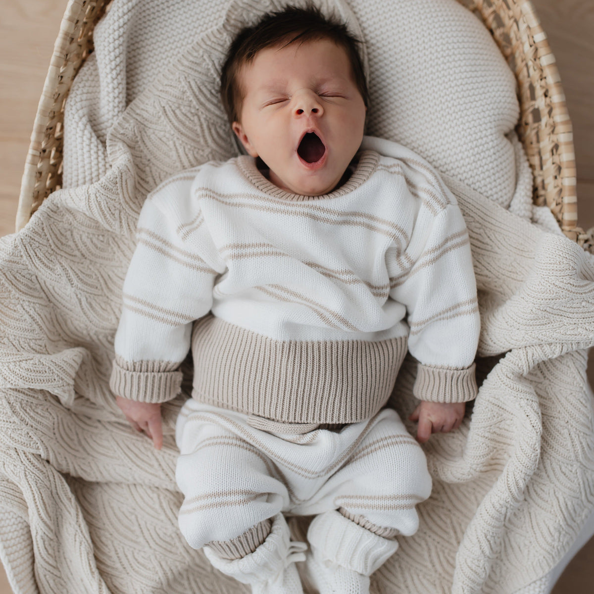 Een vredige pasgeboren baby gaapt terwijl hij comfortabel in een geweven wiegje ligt, gewikkeld in een zachte 100% Yumi Baby gebreide broek Sandy Stripe deken.