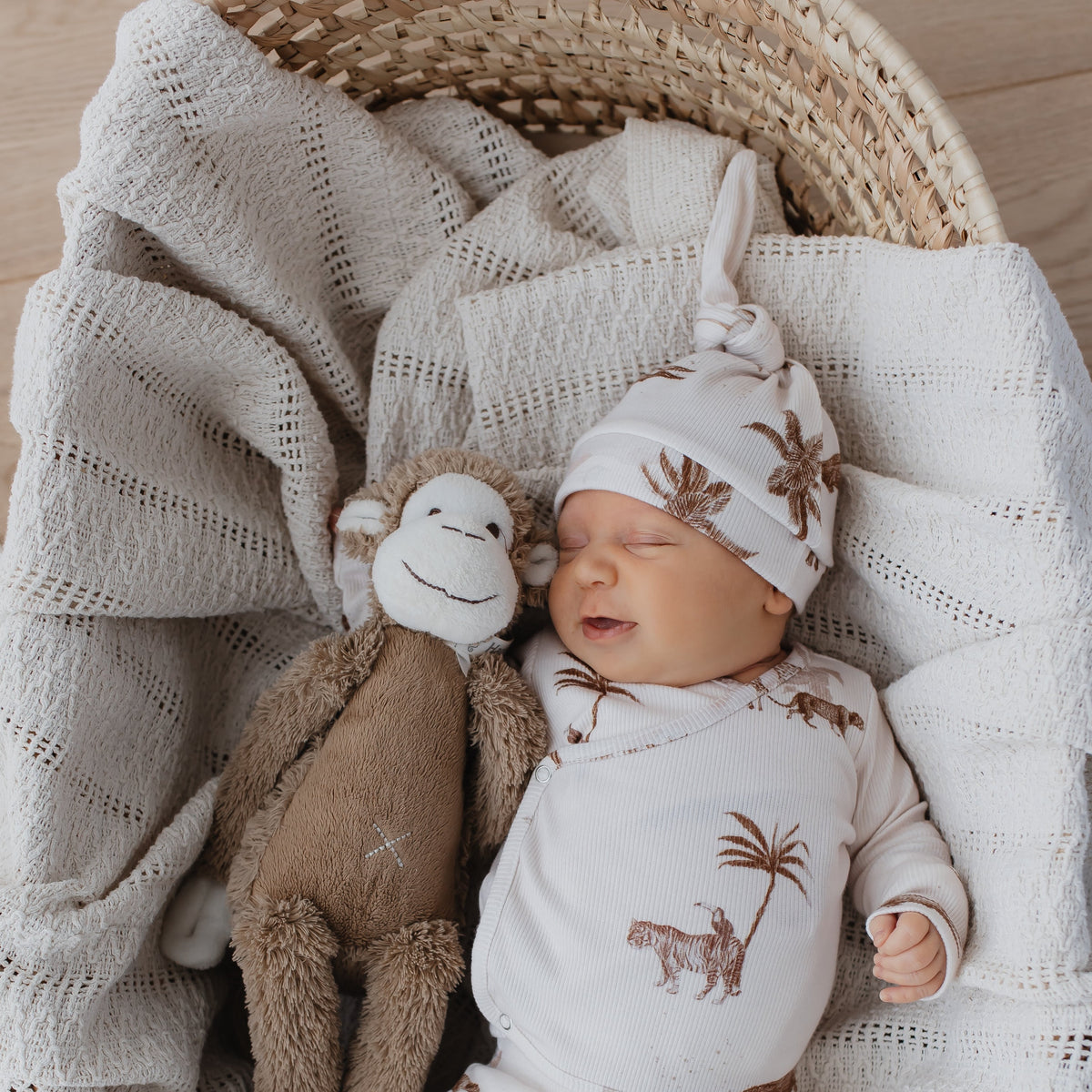 Een vredig slapende pasgeborene, knus gewikkeld in een Yumi Baby Muts Vintage Jungle Taupe-outfit, nestelt zich gezellig naast een pluchen aapvriendje in een rieten mand, gedrapeerd met een zachte, gestructureerde deken.
