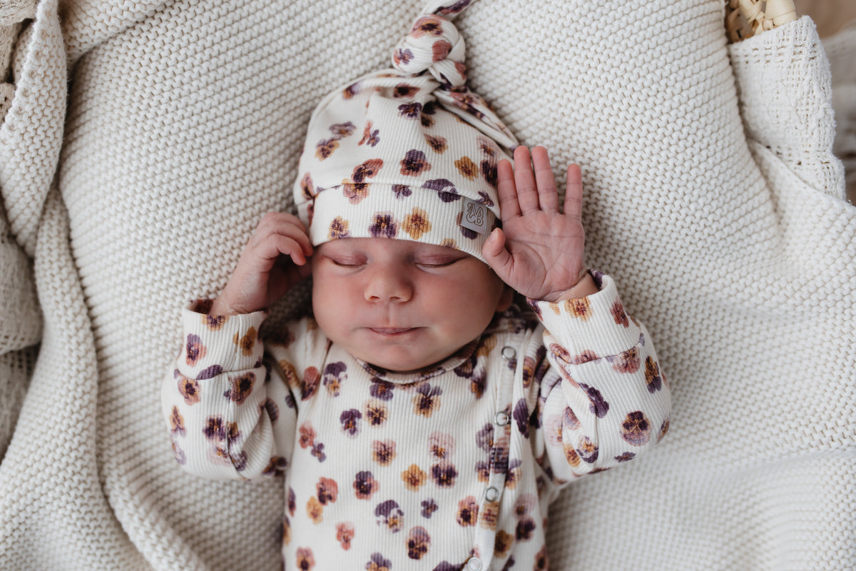 Een vredige pasgeboren baby gekleed in een Yumi Baby Boxpakje Pretty Pansies rompertje met bijpassend mutsje, zachtjes rustend op een zacht gebreide deken.