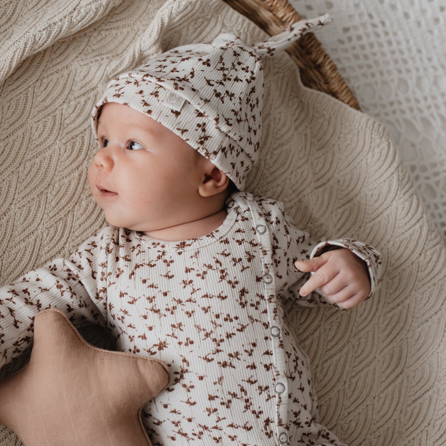 Een nieuwsgierige baby gekleed in een rompertje met patroon gemaakt van bestendige materialen en een bijpassende Yumi Baby pasgeboren Muts Brown Leafs, comfortabel liggend met een stervormig speeltje, aandachtig starend.