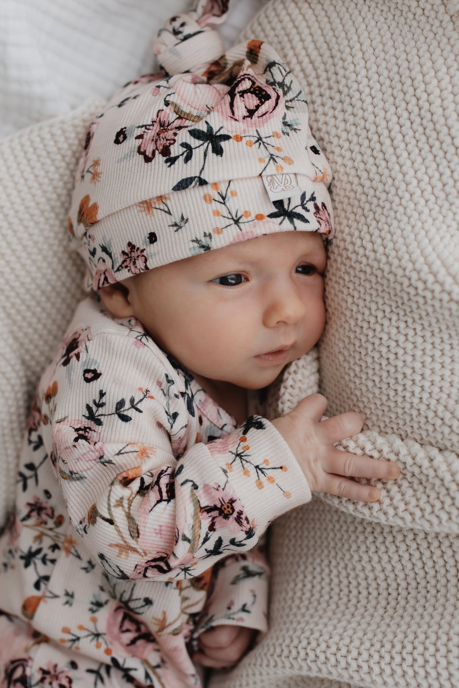 Een serene baby gekleed in een Boxpakje Blamy met bloemenmotief van Yumi Baby met een bijpassend mutsje, gezellig genesteld op een crèmekleurige deken met textuur.