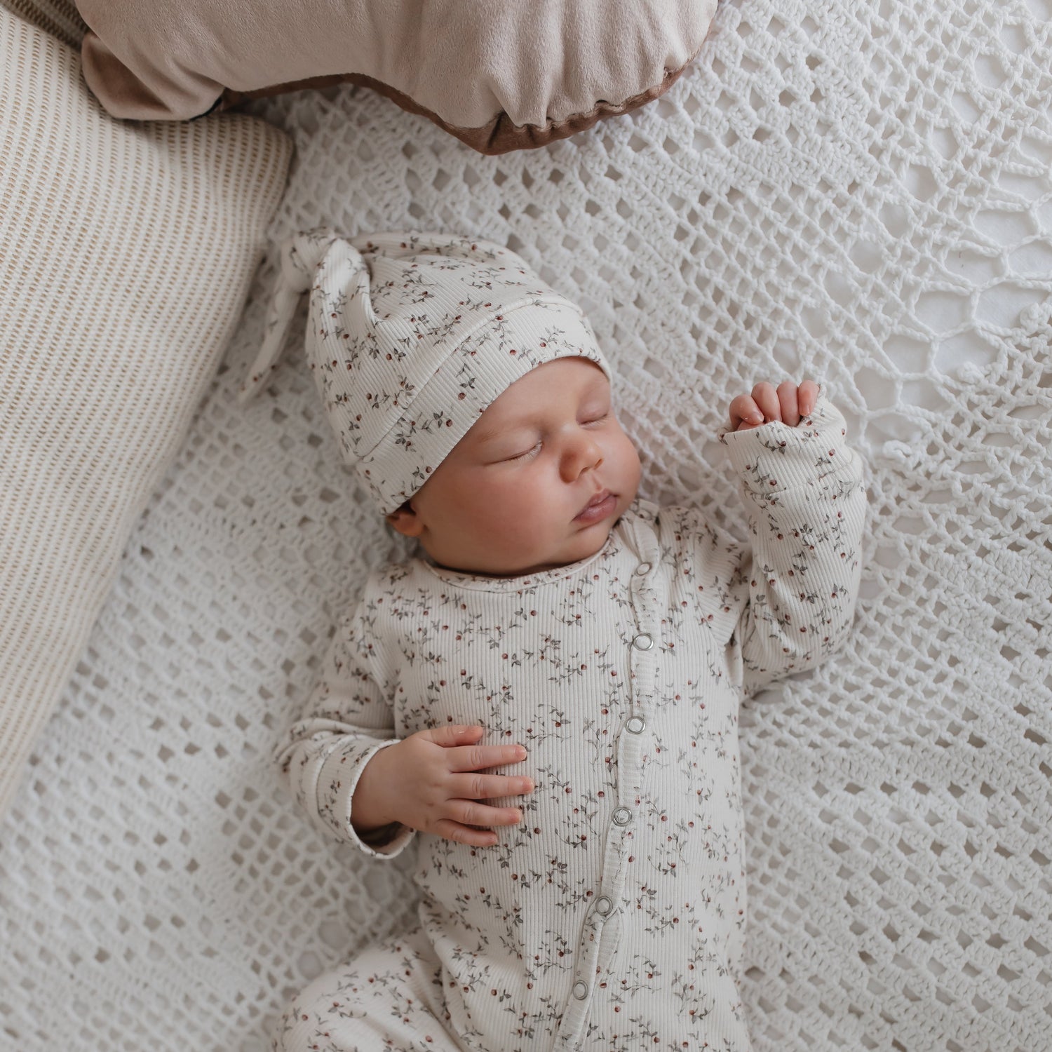 Een vredige pasgeborene slaapt heerlijk op een structuurdeken, draagt een knus rompertje met patroon en een bijpassende geknoopte Yumi Baby Muts Appeltakken.
