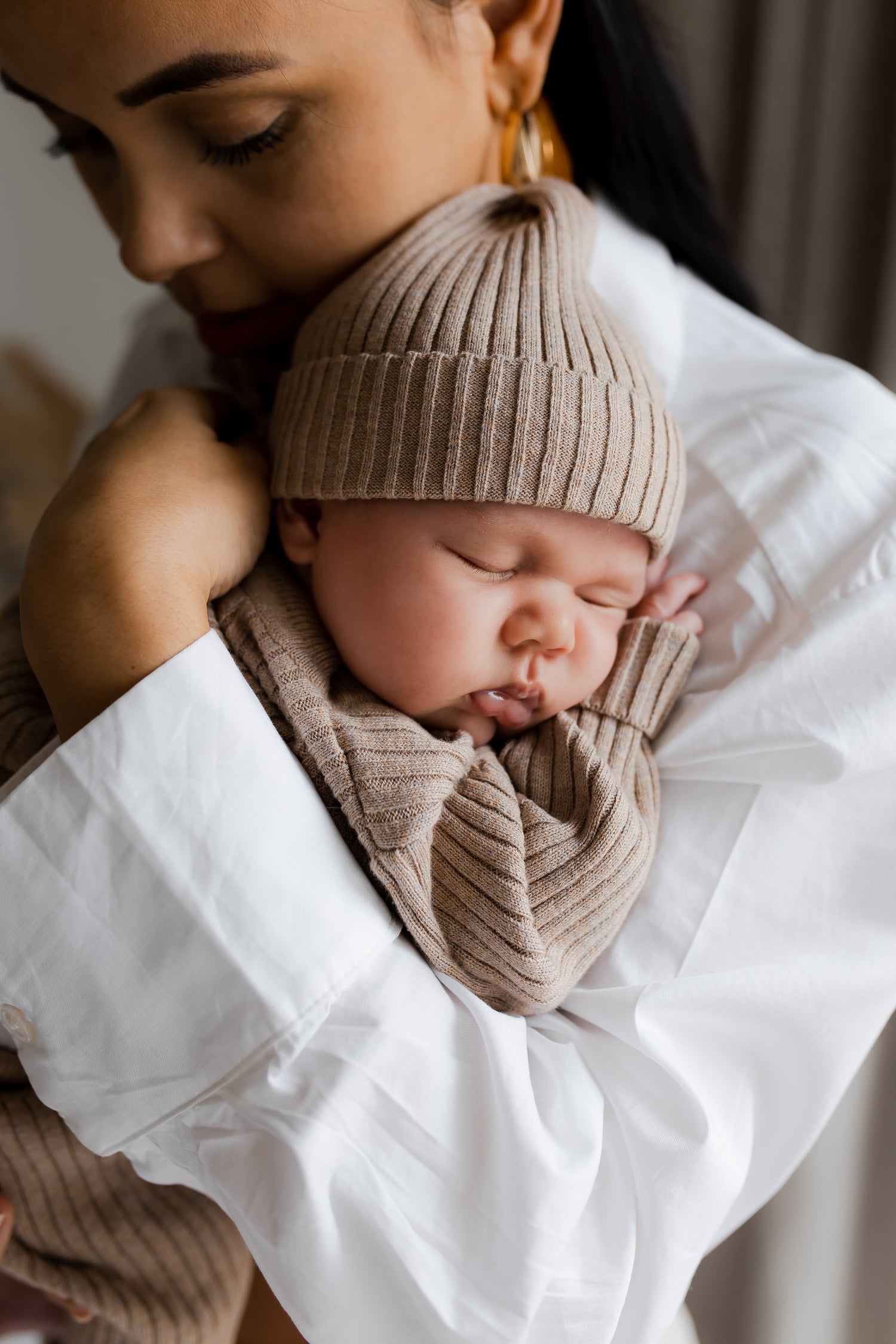 Een sereen moment waarop een moeder haar slapende pasgeboren baby teder omhelst, gekleed in een bijpassende gebreide muts Cacao Cutie muts en omslagdoek van 100% katoen, tegen haar schouder, gehuld in de warmte van Yumi Baby.