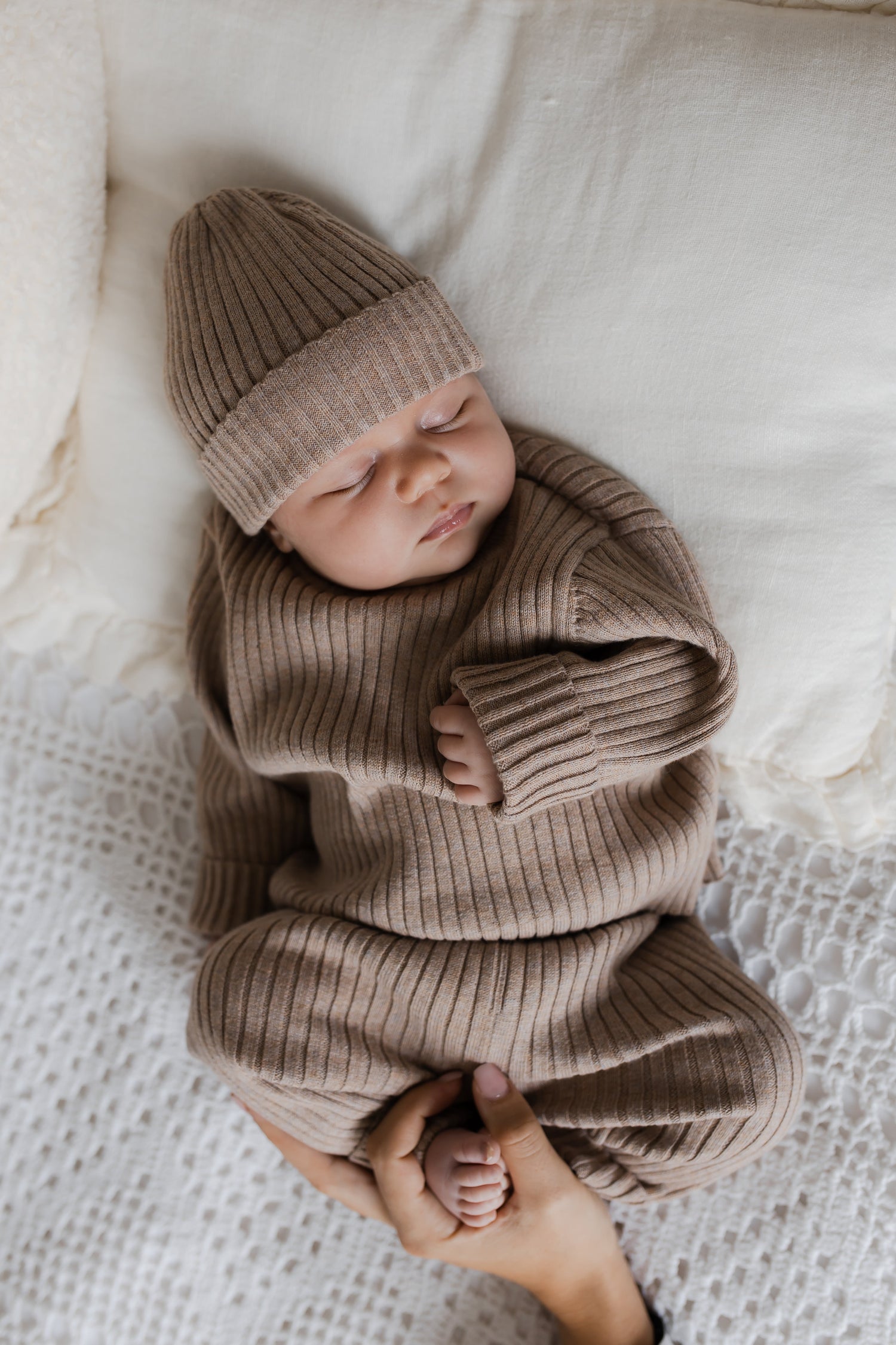 Een vredig kind gekleed in een knus Yumi Baby Knitted broekje Cacao Cutie-outfit met een bijpassende muts, slaapt comfortabel op een zacht, crèmekleurig textieloppervlak, met een zachte wieg van de hand