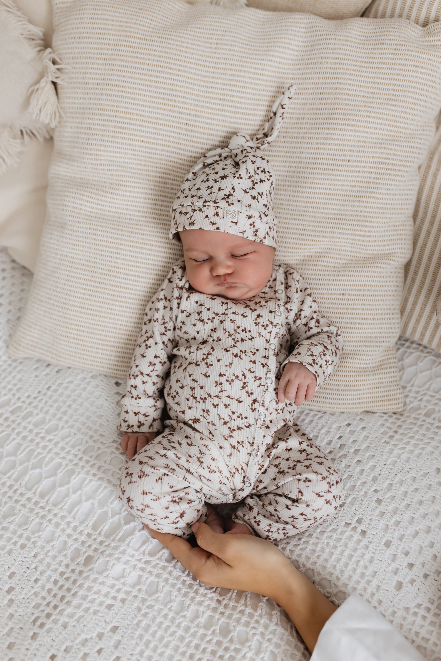 Een vredige pasgeboren baby, gekleed in een schattig Boxpakje Brown Leafs van hoogwaardig katoen en bijpassend mutsje, wegdommelend terwijl hij comfortabel tussen de zachte kussens op een knus bed ligt. Merk: Yumibaby.