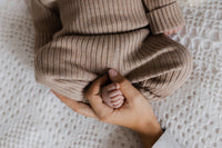 Een vredig kind gekleed in een knus Yumi Baby Knitted broekje Cacao Cutie-outfit met een bijpassende muts, slaapt comfortabel op een zacht, crèmekleurig textieloppervlak, met een zachte wieg van de hand