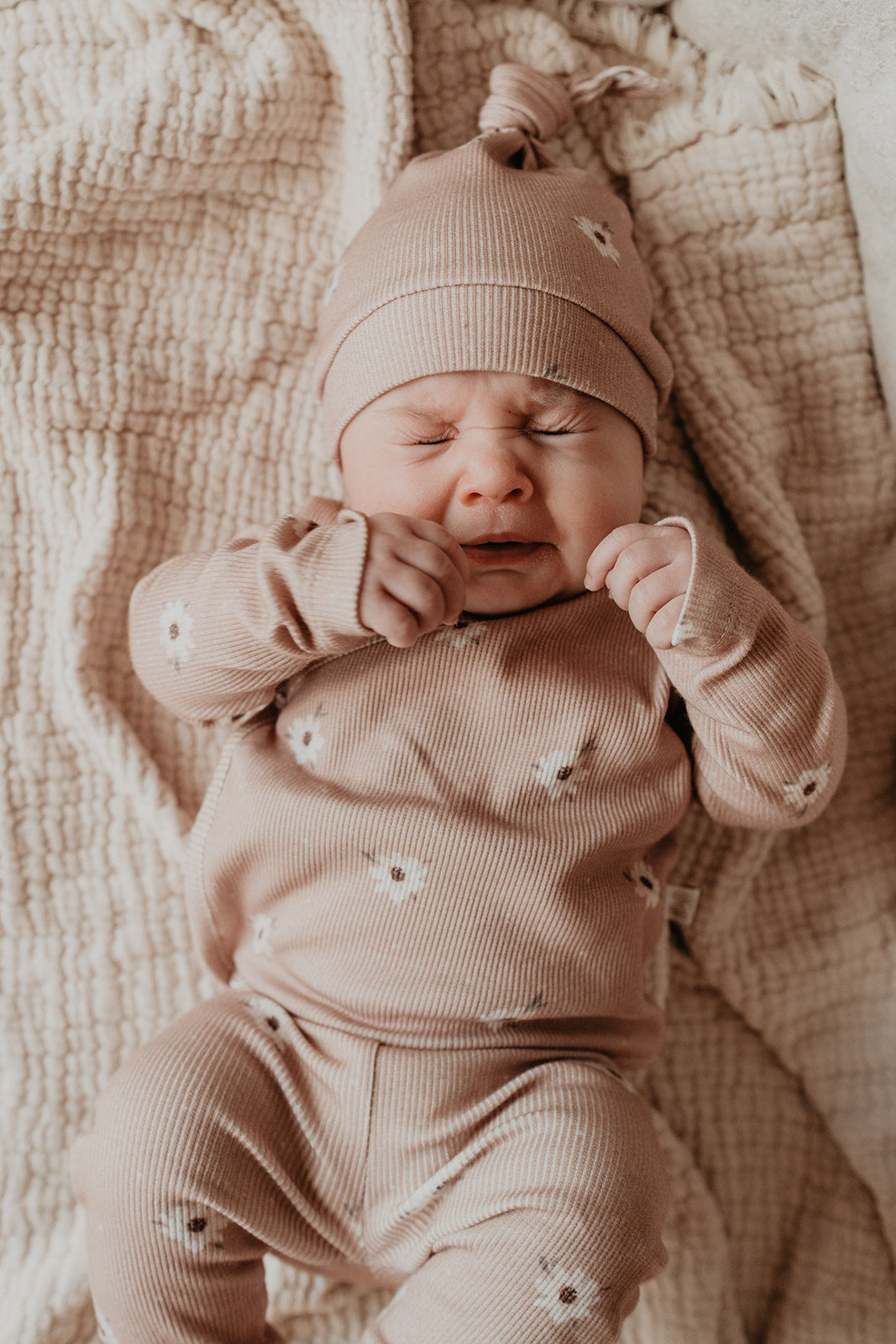 Een vredige pasgeboren baby, gekleed in een schattig, milieuvriendelijk broekje Zoë met stippen onesie met sterrenmotief en bijpassende muts, rekt zich een beetje uit of geeuwt, comfortabel liggend op een zachte, getextureerde Yumi Baby.