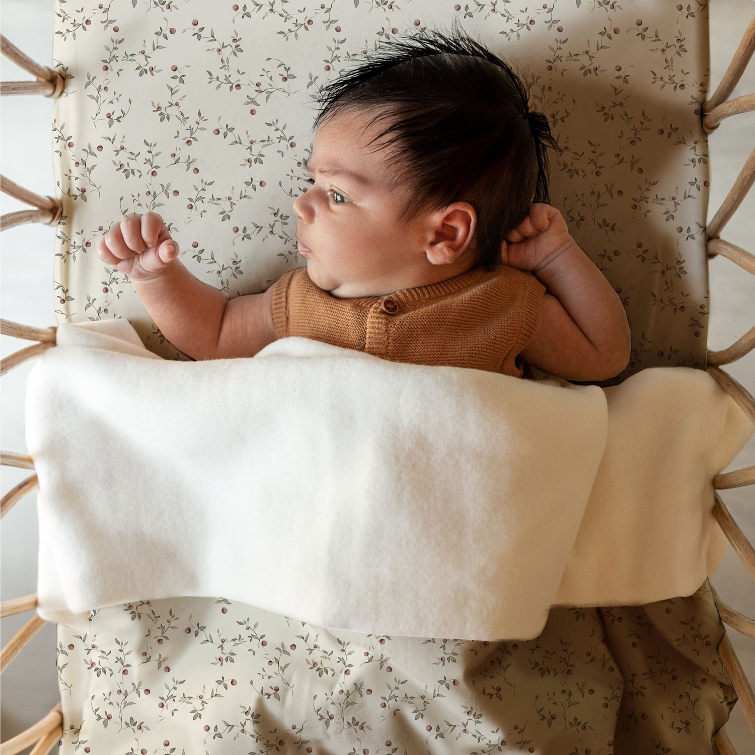 Een vredige pasgeborene met plukjes donker haar ligt op een comfortabele Yumi Baby Ledikantdeken Appeltakken, knus in een wiegje in boho-stijl, nieuwsgierig naar de wereld om zich heen kijkend.