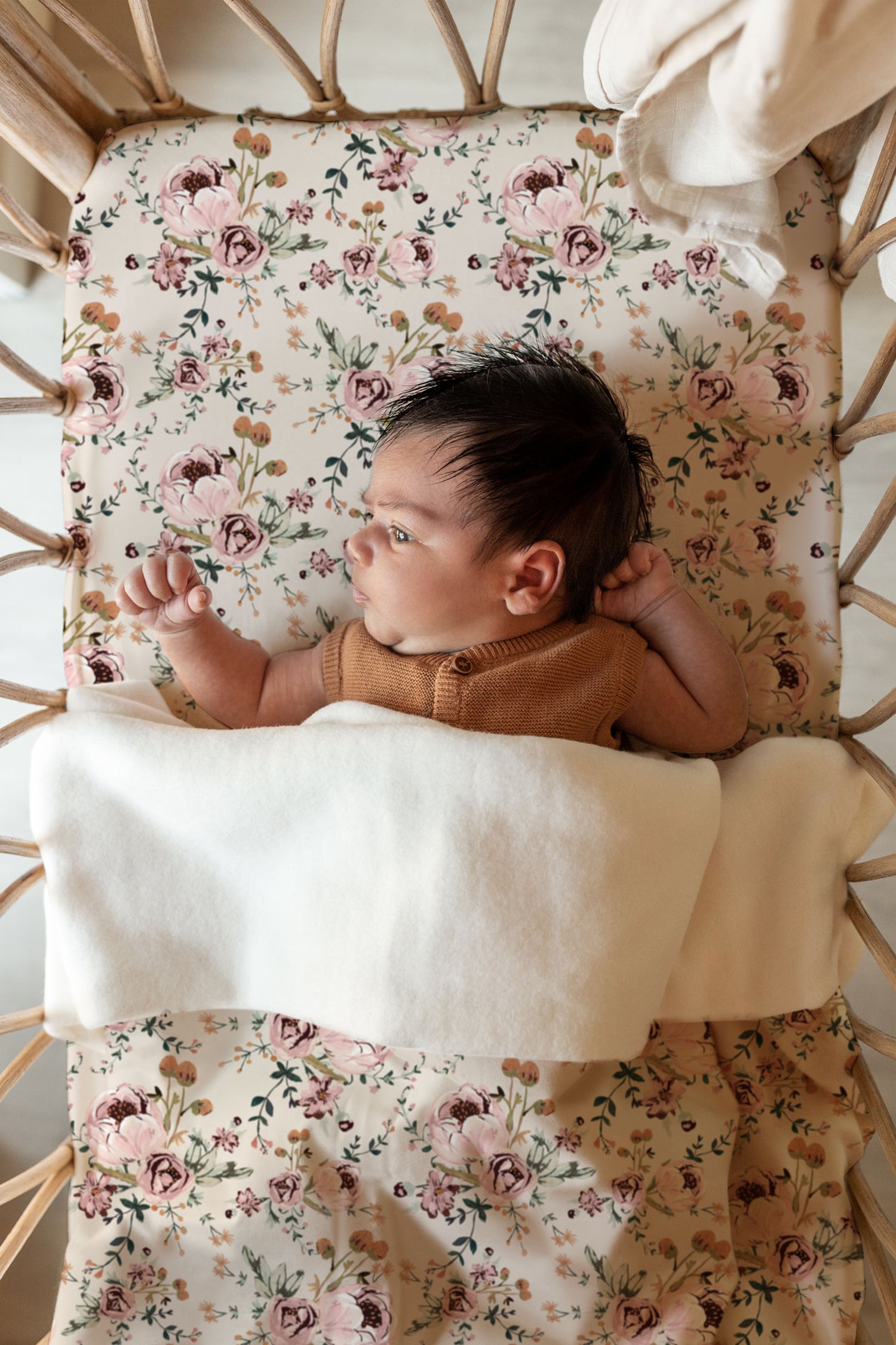 Een pasgeboren baby met donker haar, ingebakerd in een Ledikantlaken Blamy-deken van Yumi Baby, liggend in een wieg met een gebloemd matrasje, nieuwsgierig opzij starend.