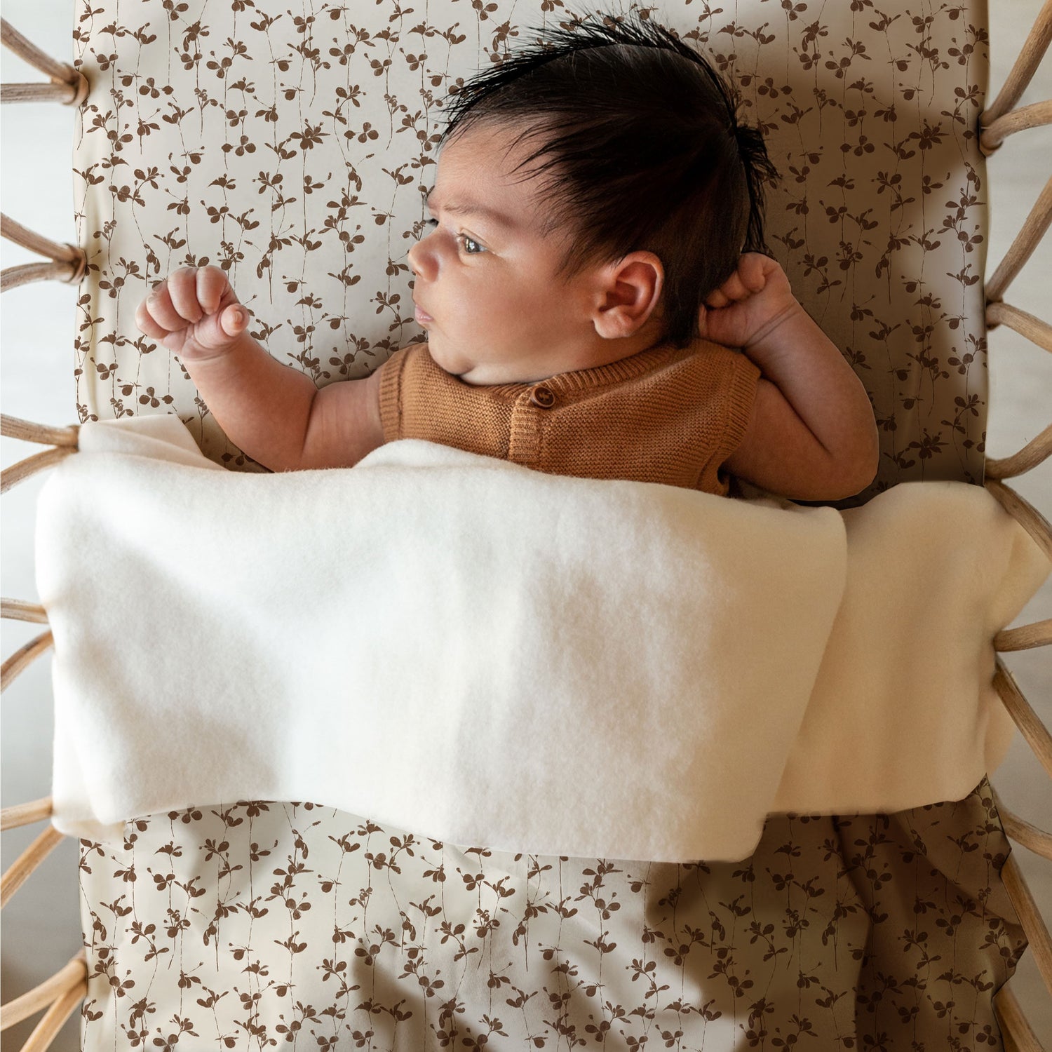 Een serene baby, gekleed in een knus bruin kledingstuk, ligt op een comfortabel oppervlak met een patroon en staart contemplatief naar de zijkant onder zachte Yumi Baby Wiegdeken Brown Leafs.
