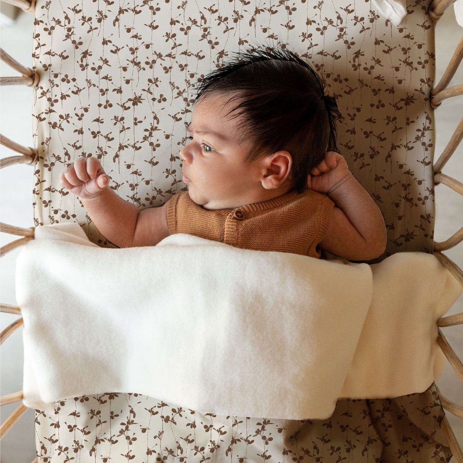 Een tevreden baby met een volle haardos ligt kalm in een wieg, gezellig ingewikkeld in een zachte witte deken, geproduceerd Ledikanthoeslaken Brown Leafs van Yumi Baby.