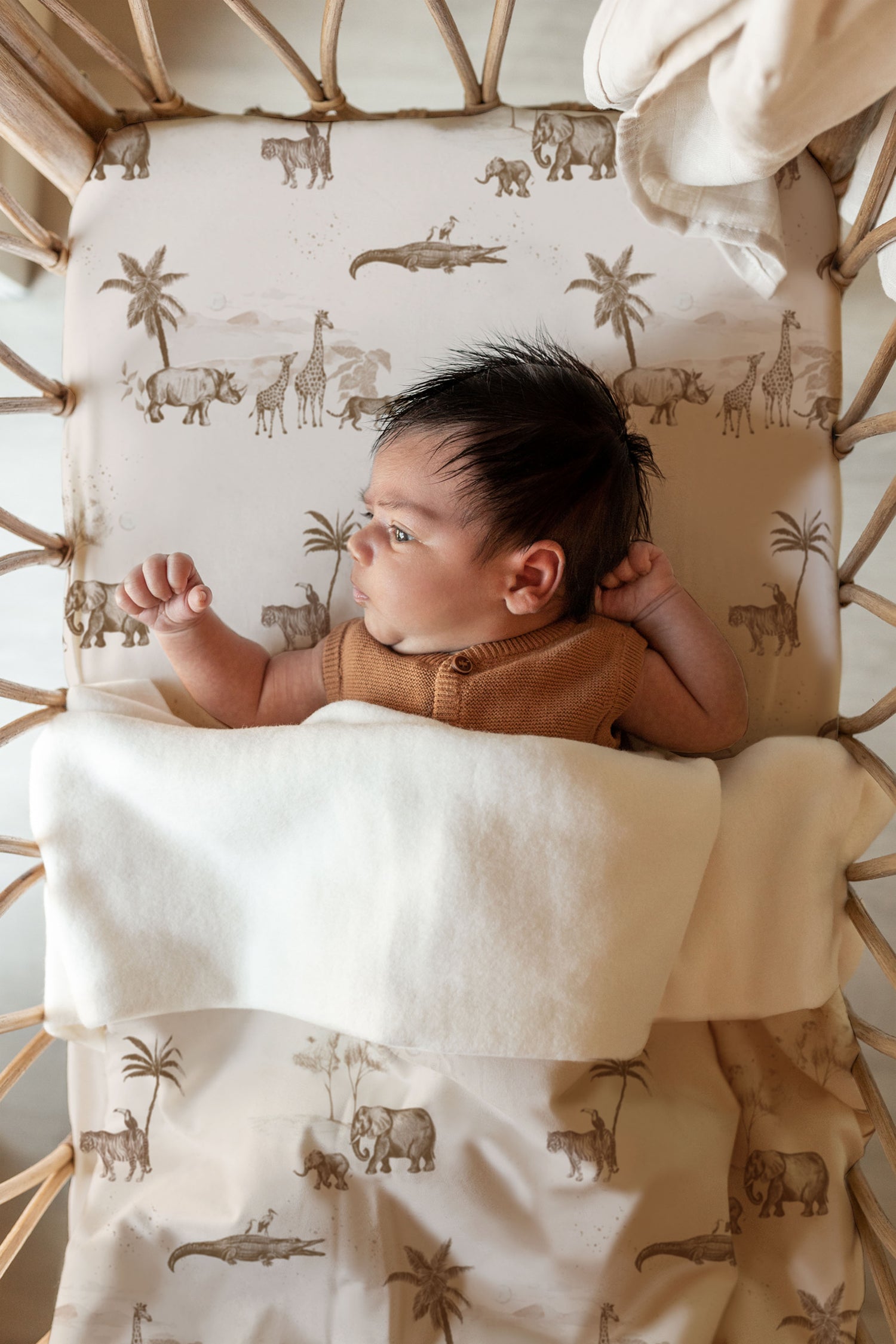 Een pasgeboren baby met een bos donker haar ligt gezellig ingebakerd in een Yumi Baby Wiegdeken Vintage Jungle Taupe deken en kijkt nieuwsgierig tegen een achtergrond met safarithema.