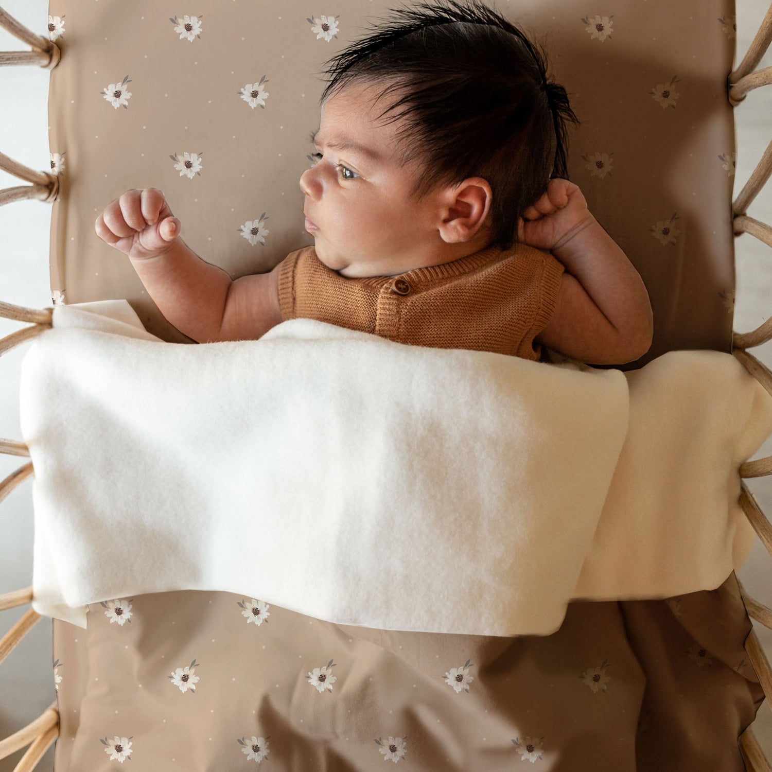 Een pasgeboren baby met een volle bos haar, gekleed in een gezellige bruine outfit, ligt op een beige kussen met patroon onder een zachte witte Yumi Baby Oeko Tex katoenen deken, met een bedachtzame uitdrukking.