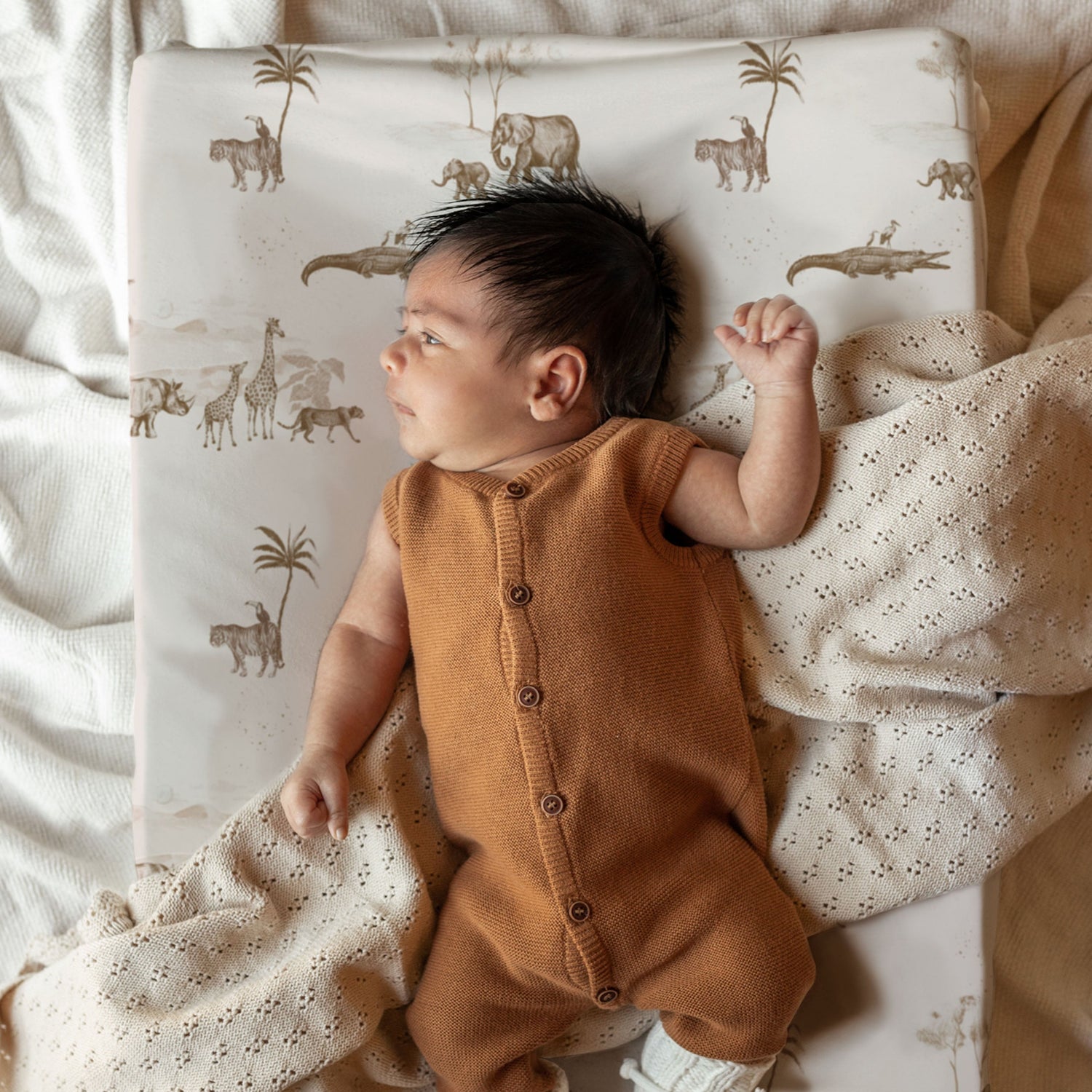 Een vredige pasgeboren baby gekleed in een knus bruin rompertje, liggend op een bed met lakens met handgemaakte prints, gedeeltelijk bedekt met een zacht dekentje, droomend in slaap op het Yumi Baby Aankleedkussenhoes Vintage Jungle Taupe.