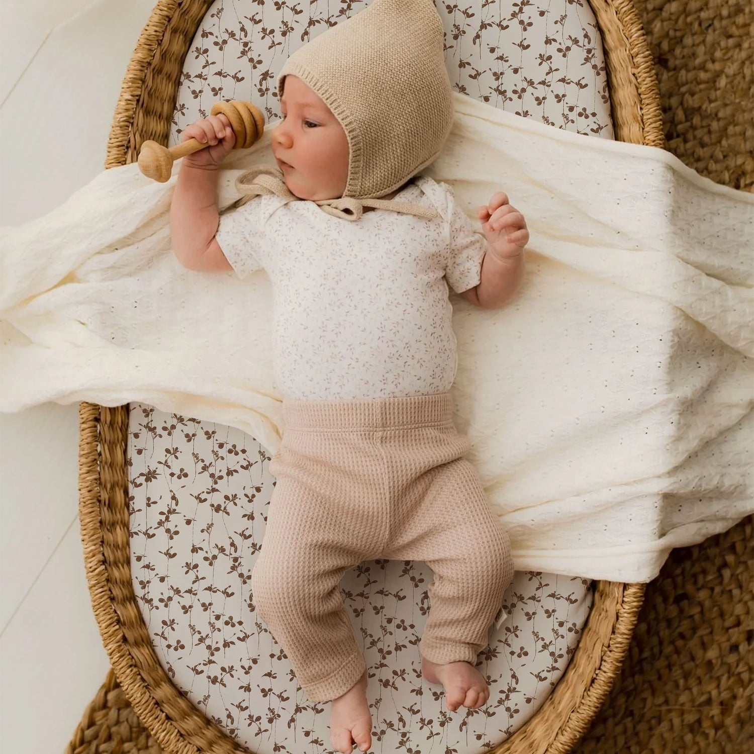 Een serene baby, gekleed in een beige gebreide muts en een bedrukt Oeko Tex katoenen rompertje, ligt ontspannen in een geweven mand, met een houten speelgoedje in de hand. Deze baby ligt heerlijk knuffelig op een Yumi Baby Wieghoeslaken Brown Leafs.