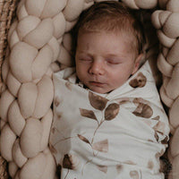Een serene pasgeborene slaapt vredig, ingebakerd in een Yumi Baby Inbakerdoek met Golden Hour-bladmotief, genesteld in een gezellig, gebreid nest.