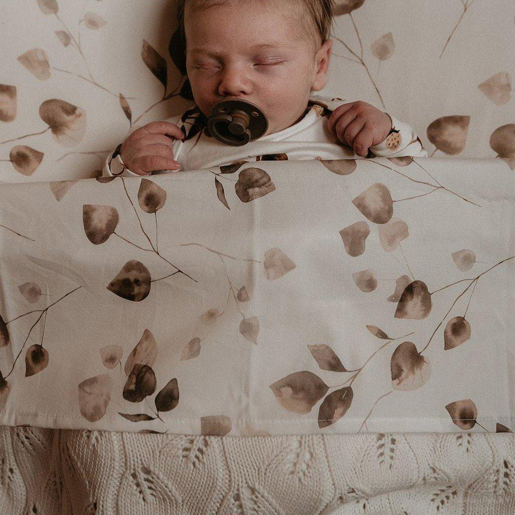 Vreedzame slaap: een baby met mollige wangen dut tevreden met een fopspeen, ingebakerd in een Yumi Baby Ledikantlaken Golden Hour-deken met een subtiel patroon.