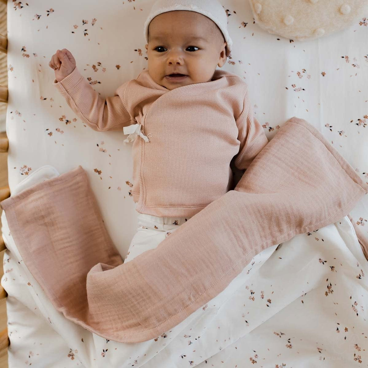 Een tevreden baby liggend op een Yumi Baby Wiegdeken deken met kersenbloesemmotief, gekleed in knusse pastelkleurige kleding en een schattige hoofdband, met een dromerige uitdrukking en een klein vuistje omhoog.