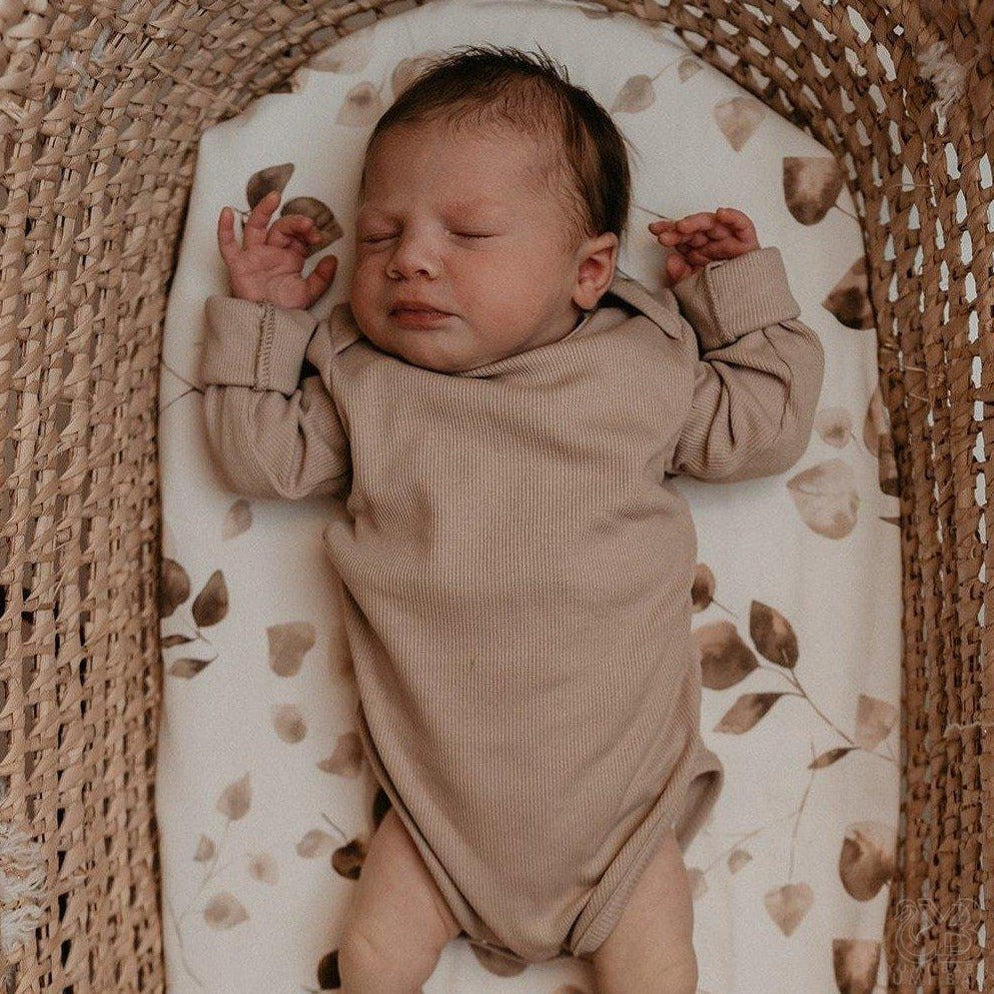 Een vredige pasgeboren baby slaapt heerlijk in een co-sleeper, gekleed in een knus beige rompertje van Oeko Tex-katoen, omringd door delicate gedroogde bladeren en Wieghoeslaken Golden Hour-lakentjes van Yumi Baby.