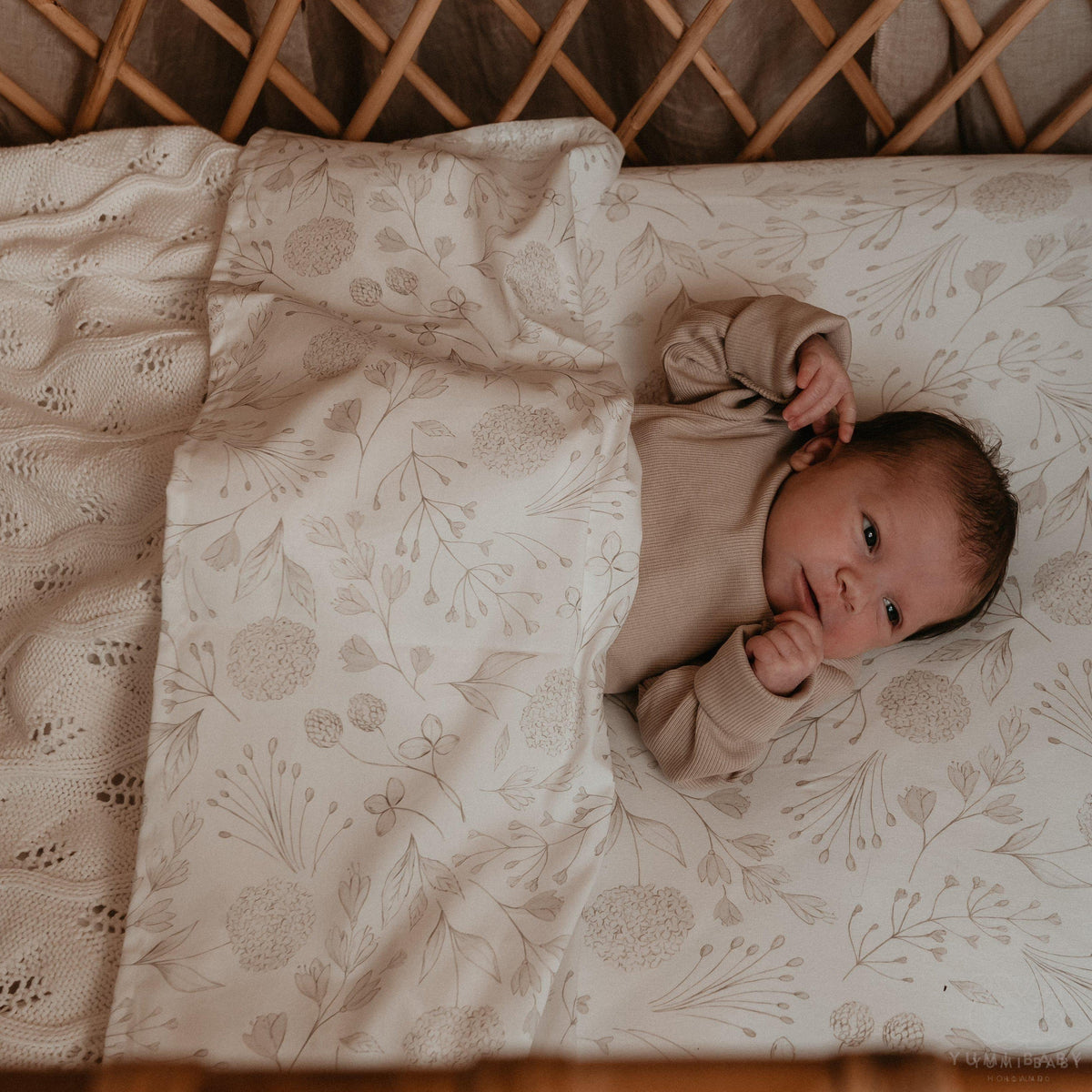 Een pasgeboren baby met waakzame ogen ligt gezellig ingebakerd in een Yumi Baby Wieglaken Sprookjesachtige crèmekleurige deken met een delicaat bloemmotief, rustend in een natuurlijk houten wiegje.