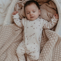 Een vredig slapende pasgeboren baby, gekleed in een witte Yumi Baby Oeko Tex katoenen onesie met delicate paarse bloemmotieven, rustend op een crèmekleurige deken met een gegolfde rand.